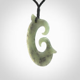 This picture shows a large hand-carved contemporary koru pendant. It is carved from New Zealand Kokopu jade and is suspended from a hand-plaited, black cord.